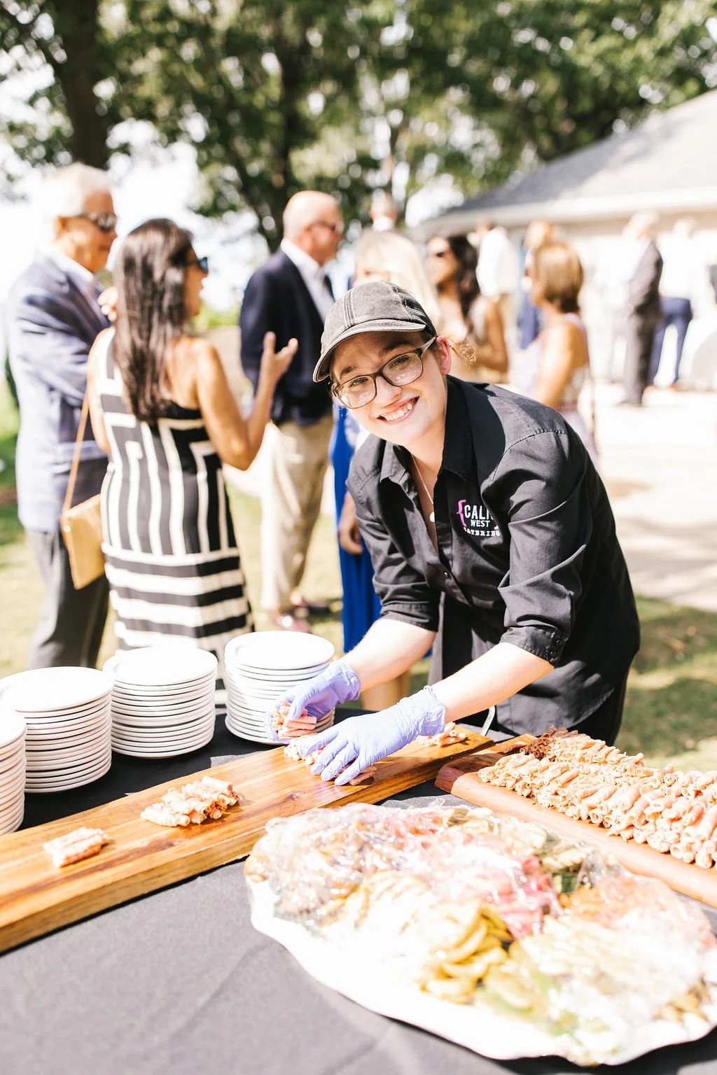 A smiling woman wearing glasses, a black shirt, purple gloves, and a hat, preparing food at an outdoor event with guests in the background.