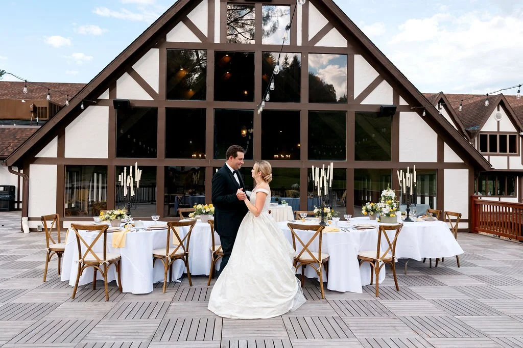 A bride and groom dancing on an outdoor deck with a large, decorated wedding reception table in front of a large building with glass windows.