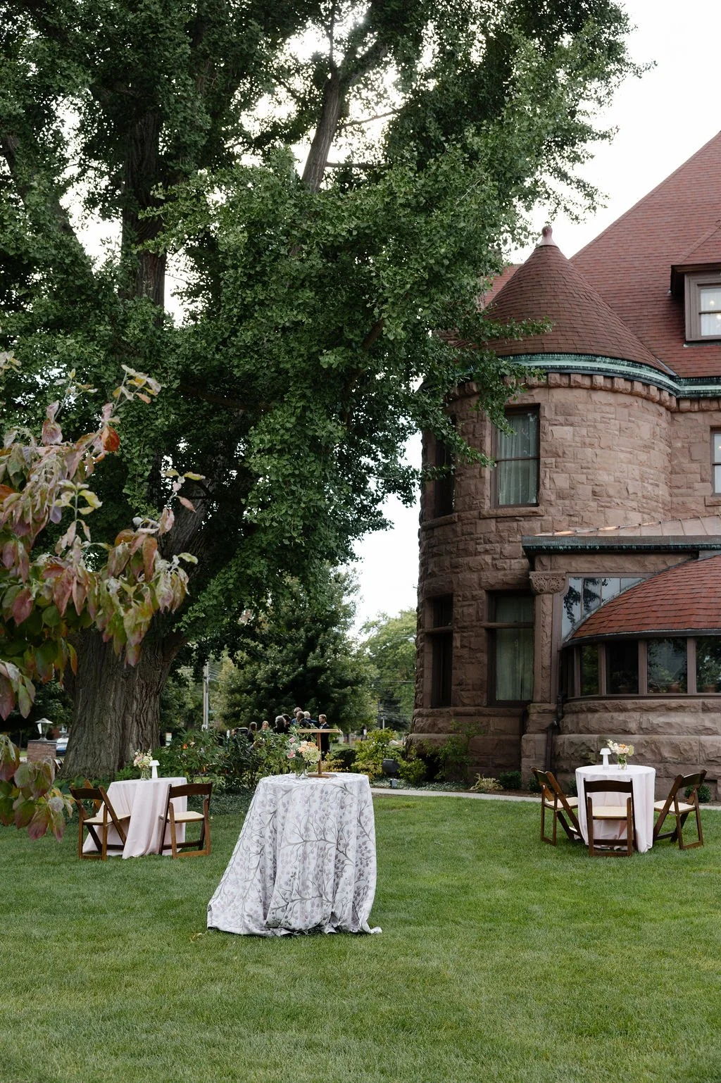 Outdoor garden scene with three tables decorated with white tablecloths and small floral arrangements, surrounded by chairs, in front of a large old stone mansion with red-tiled roof and a large tree with dense green foliage.