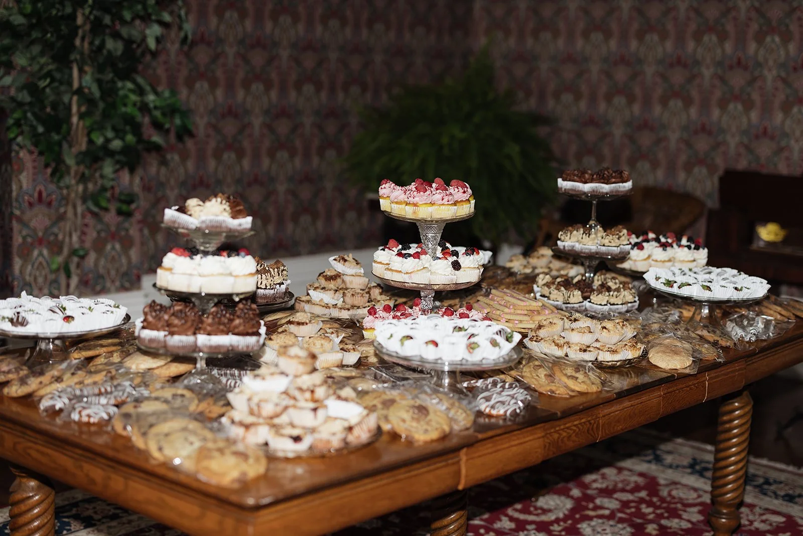 A large wooden table decorated with an assortment of holiday desserts, including cakes, cookies, and pastries, on glass cake stands and plates, with a patterned wallpaper and potted plant in the background.