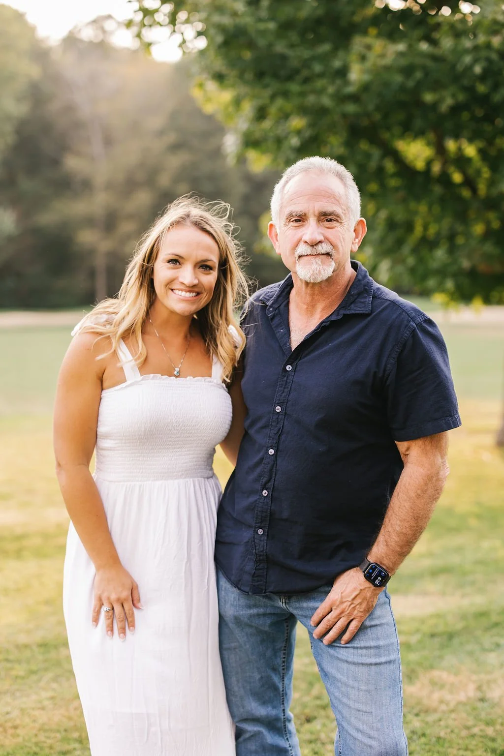 A young woman and an older man standing outdoors in a park, smiling at the camera. The woman is wearing a white dress with shoulder straps and a necklace, while the man is wearing a navy blue short-sleeve shirt, jeans, and a smartwatch on his left wr