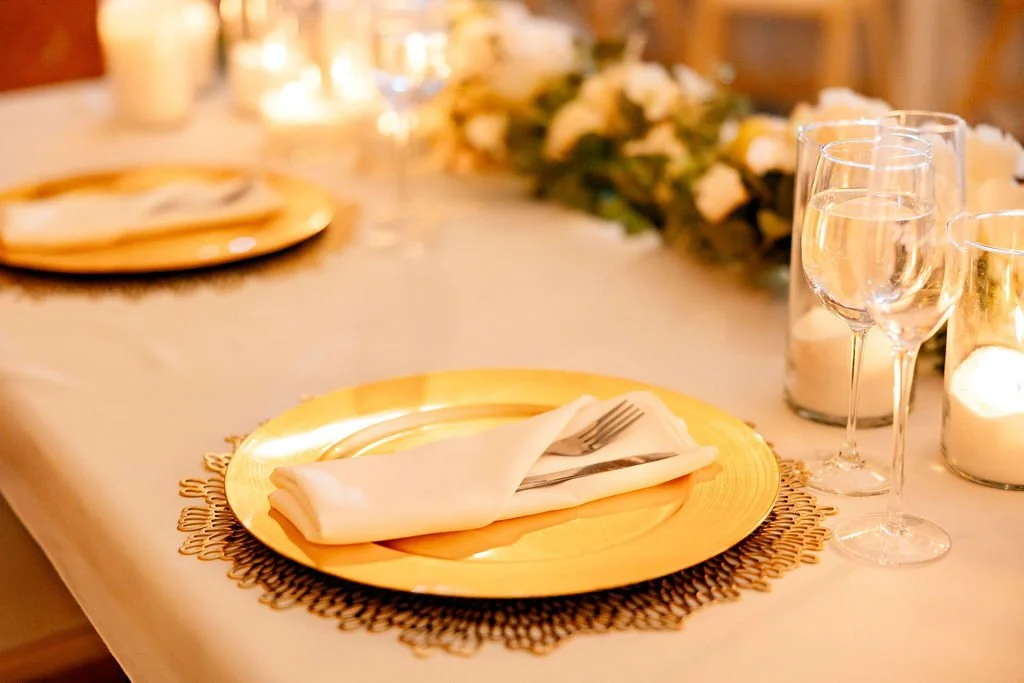 A table setting with a gold charger plate, white napkin, fork and knife, wine glasses, candles, and floral decoration in a warm, softly lit environment.