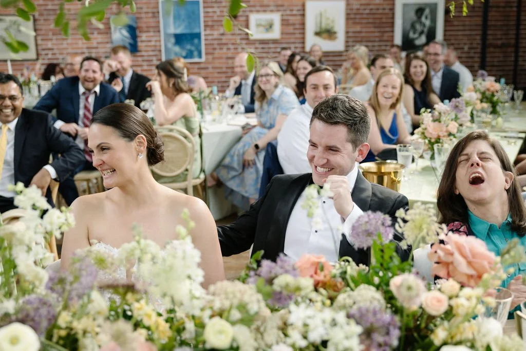 Group of people at a wedding reception, smiling and laughing, with floral decorations and seated at long tables inside a venue with brick walls and artwork.