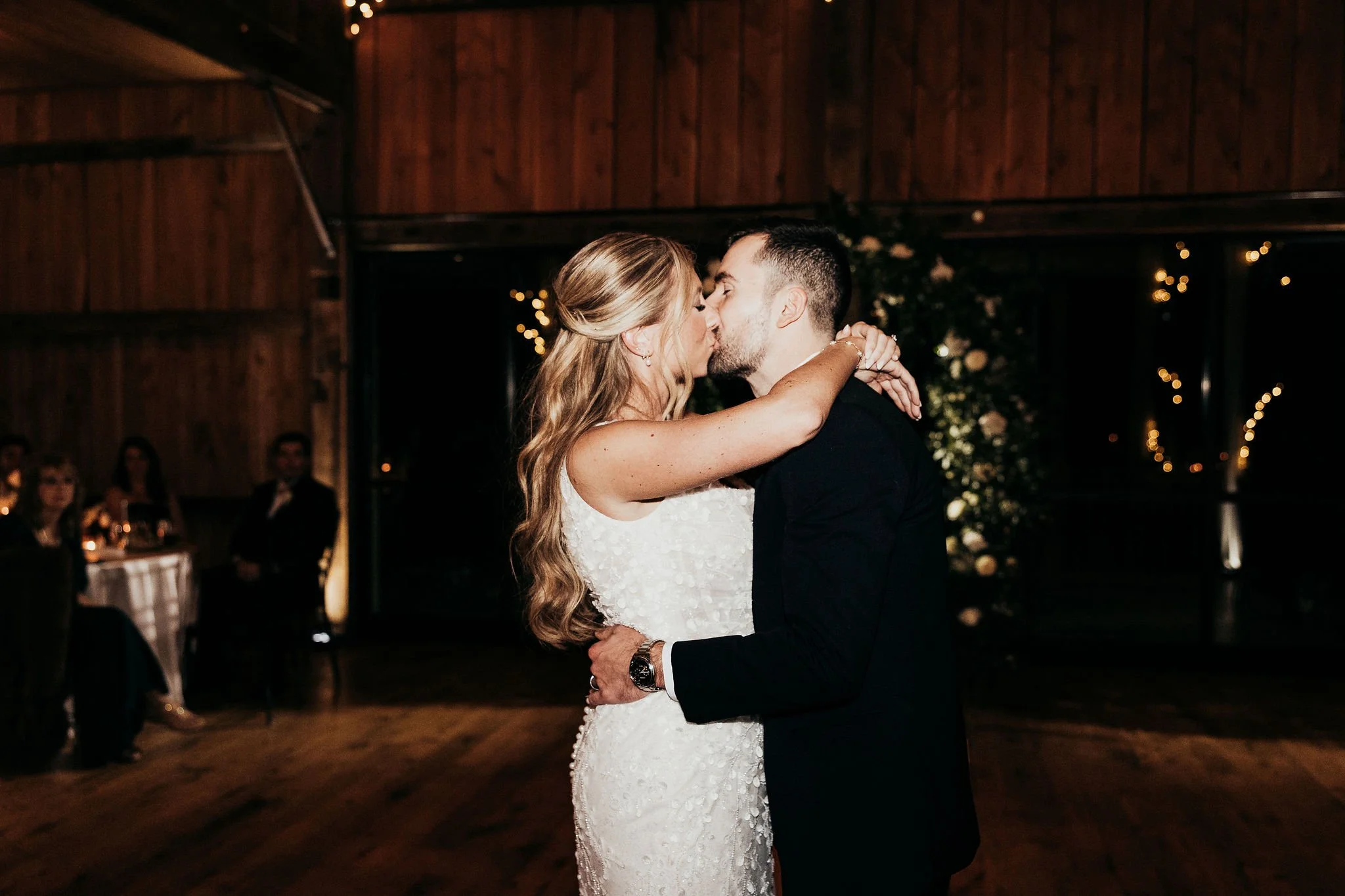 A bride and groom are dancing closely in a rustic indoor wedding reception, with guests seated at tables in the background and string lights adding warm ambiance.