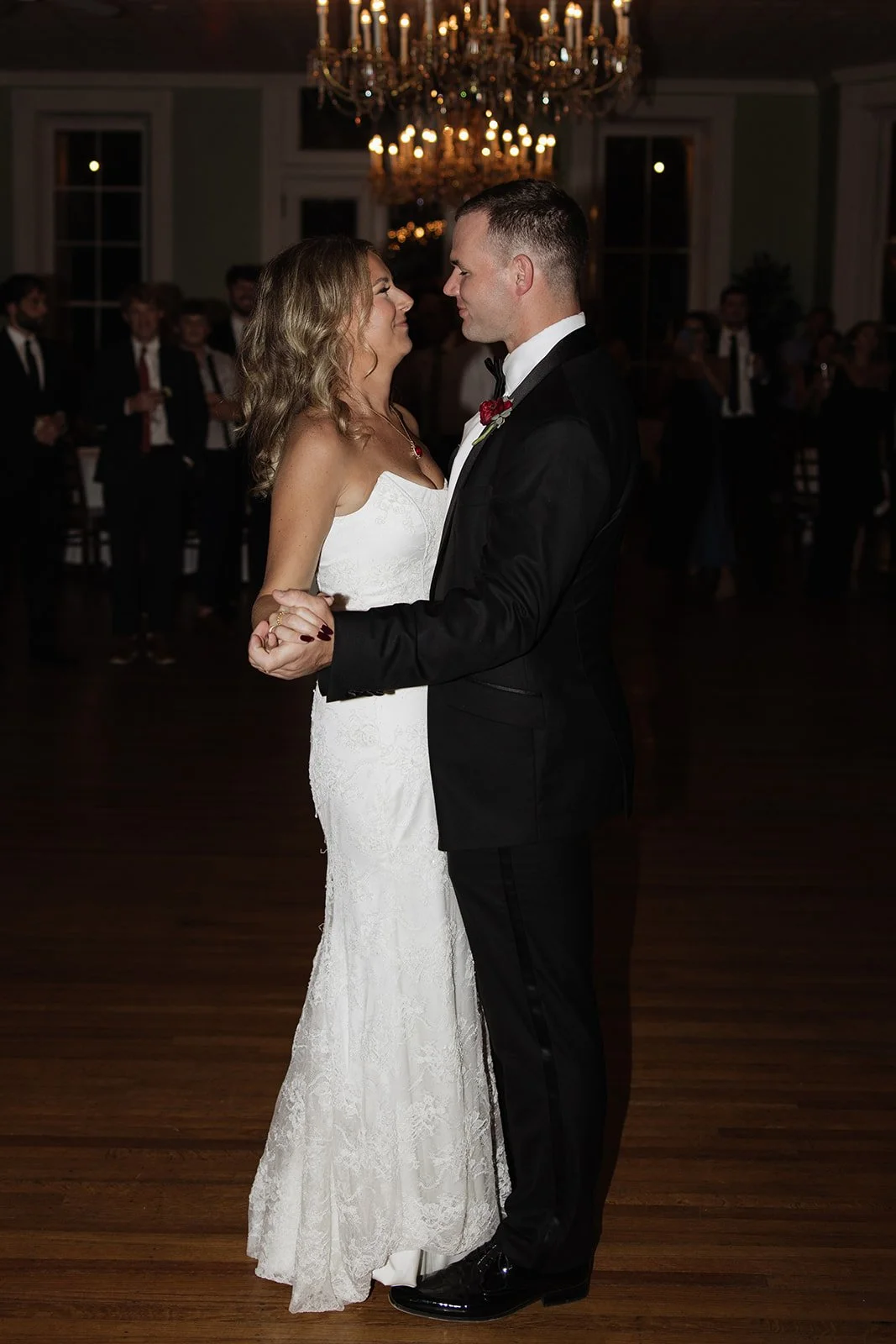 A bride and groom share a first dance in a dimly lit ballroom with chandeliers, surrounded by guests.