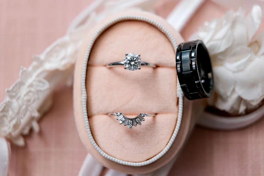 A jewelry box with two rings inside, a diamond engagement ring and a wedding band, next to a set of black rings and a lace ribbon.