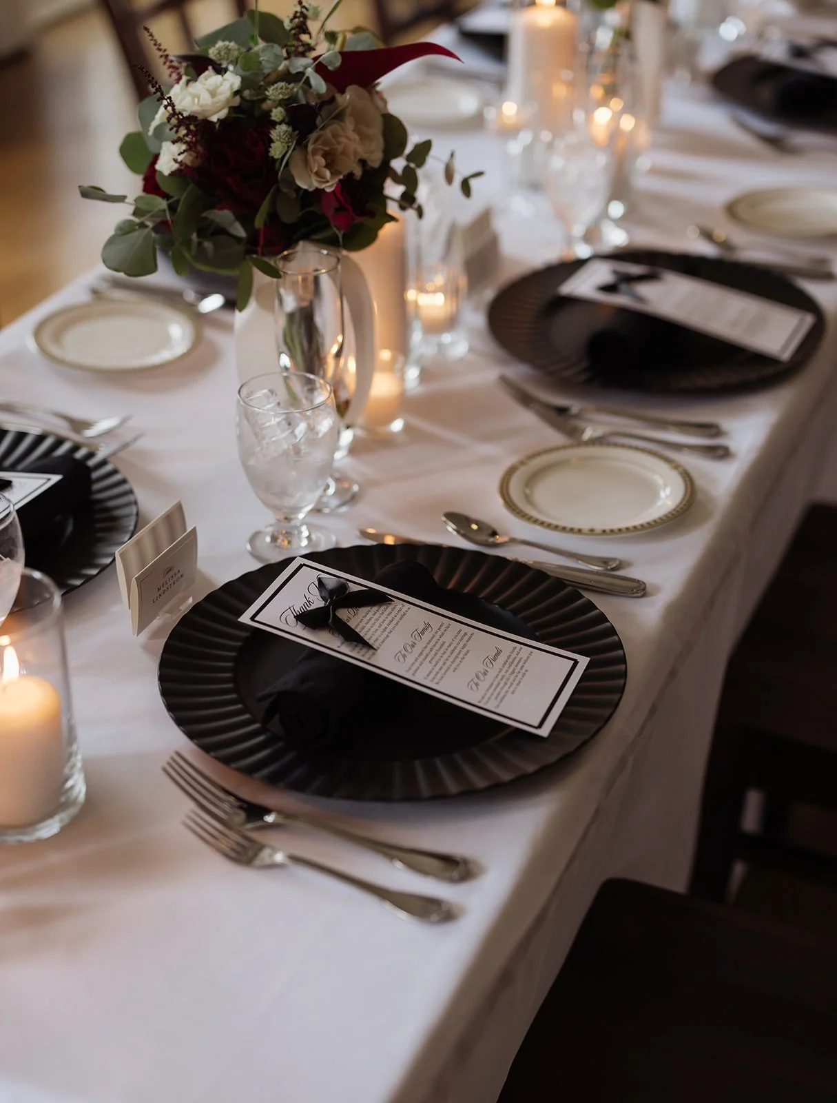 Elegant table setting with black and white plates, wine glasses with ice, silverware, a floral centerpiece with white and red flowers, candles, and a printed menu on black napkins.