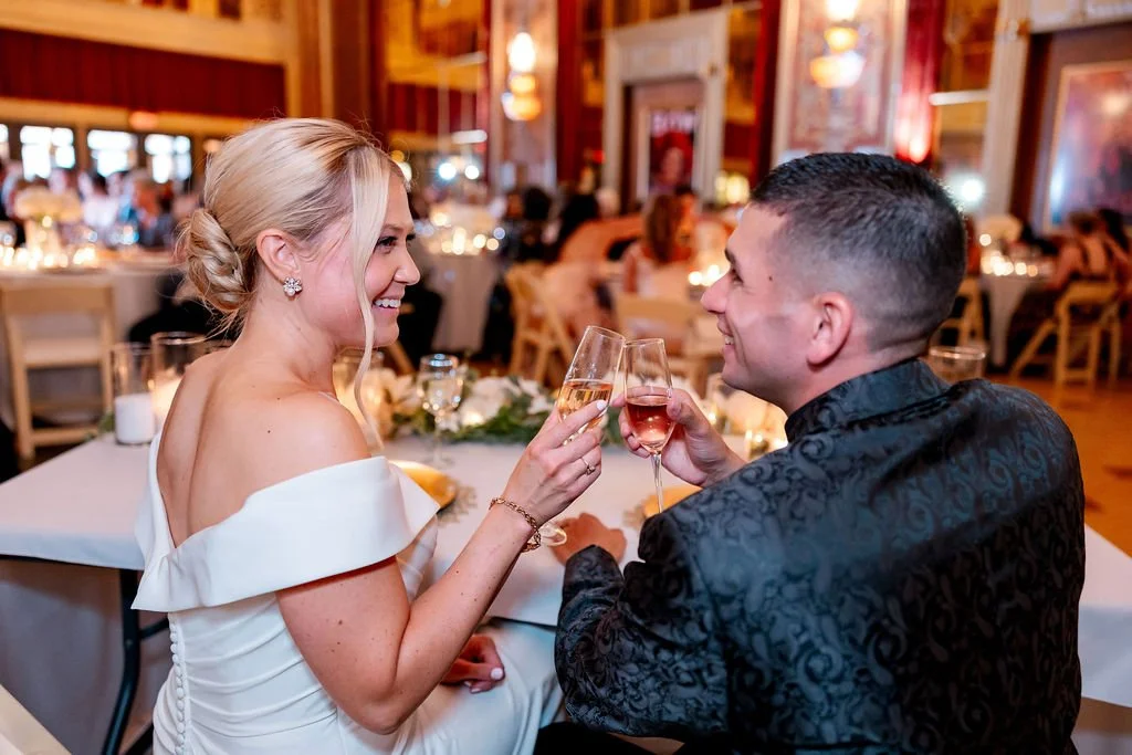 A couple at a wedding reception toasting with champagne glasses in a decorated venue with tables and guests in the background.