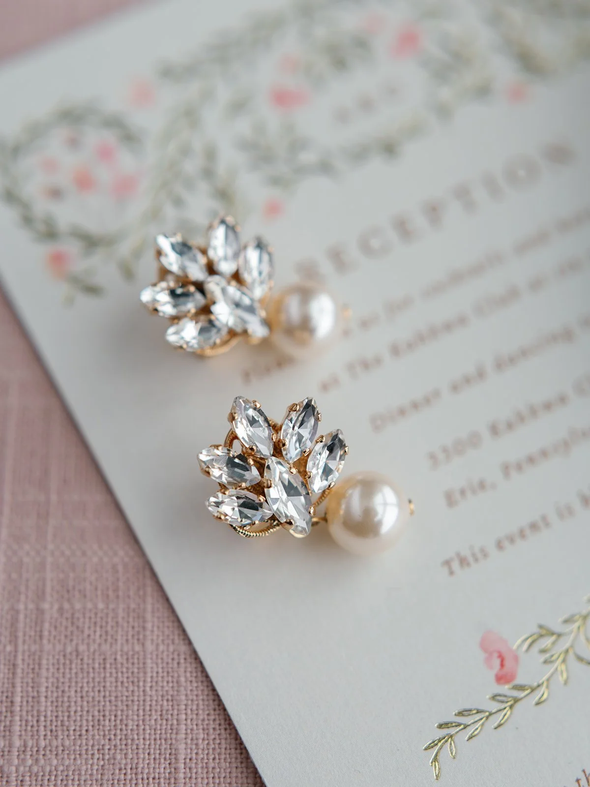 Close-up of pearl and rhinestone leaf-shaped earrings on a floral-themed wedding invitation with pink and gold accents.