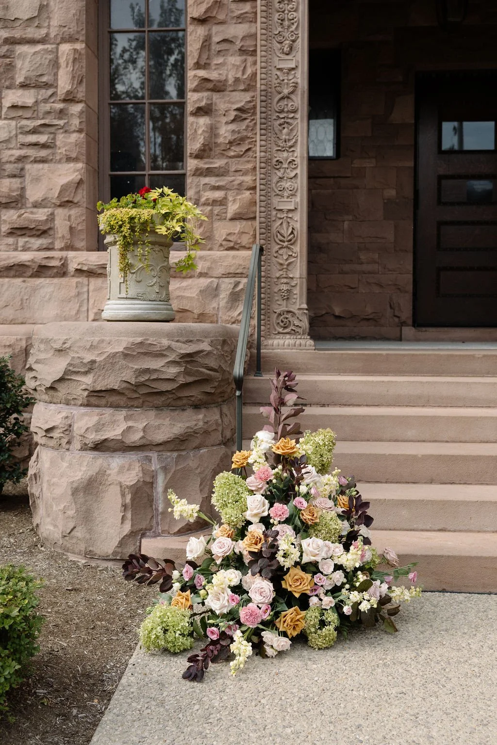 A floral arrangement on the ground near a set of stairs leading to a stone house entrance, with a decorative stone column and an urn with green and red plants on the porch.