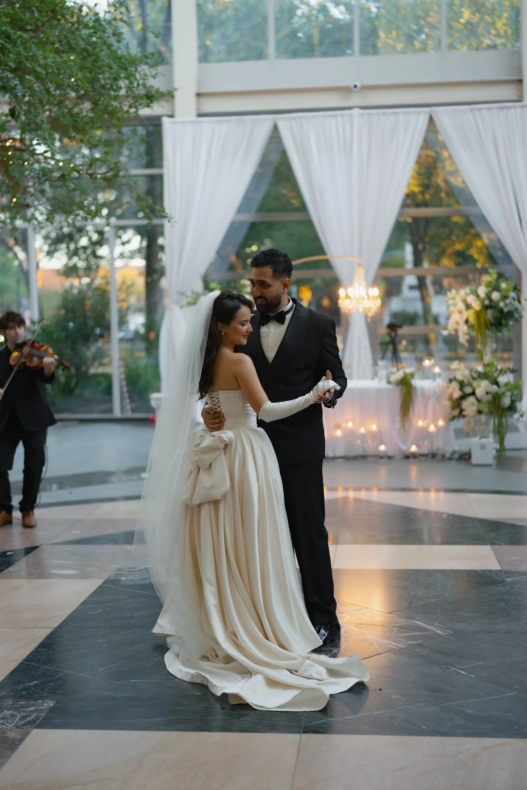 A bride and groom dancing at a wedding reception indoors with large windows and white curtains, a chandelier, floral arrangements, and a musician playing a violin in the background.