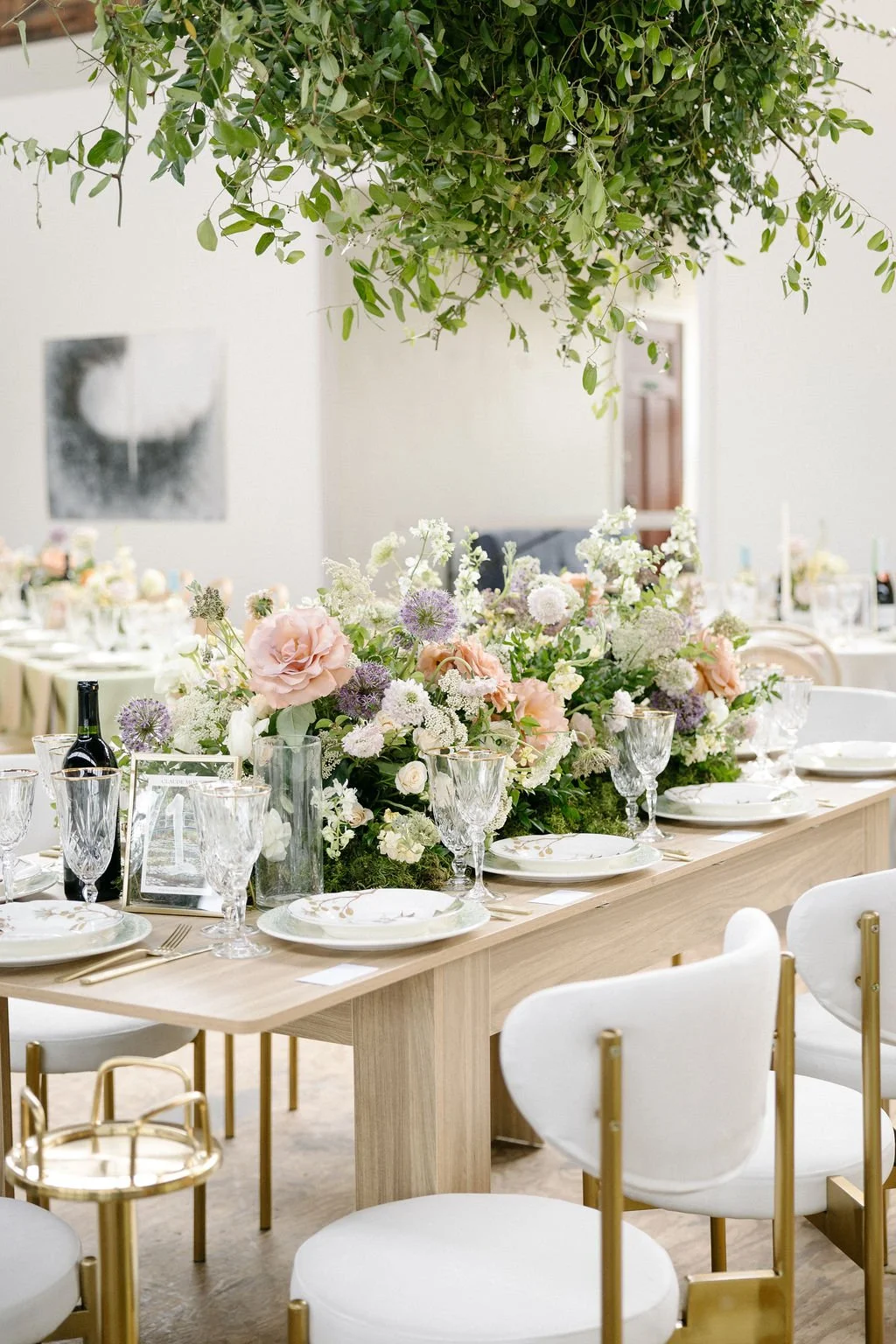 Elegant dining table decorated with a lush floral centerpiece featuring pink, white, and purple flowers, set for a formal event with crystal glassware, white plates, and gold cutlery, with white and gold chairs and a green leafy plant hanging above.