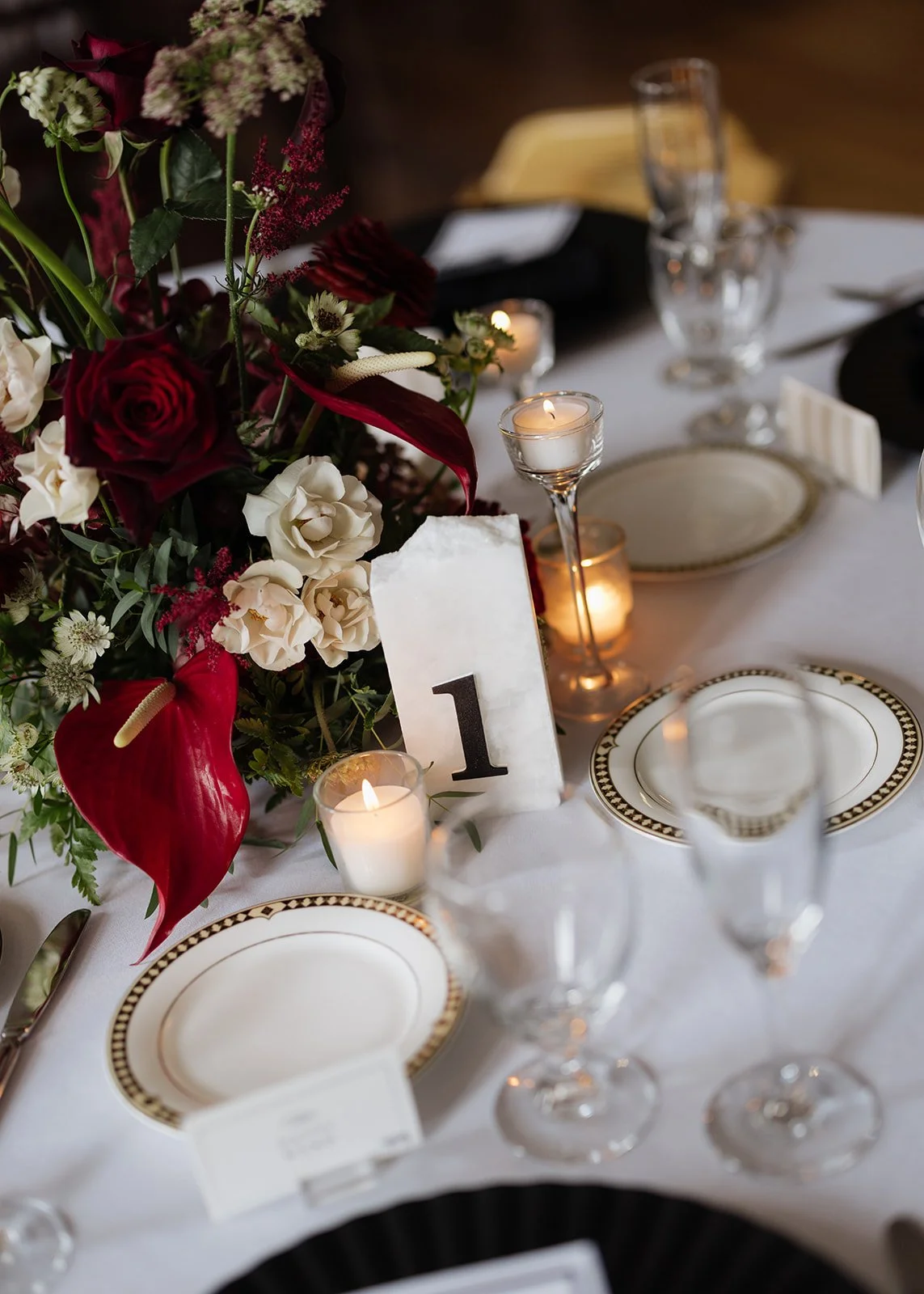 A beautifully set formal dining table with white tablecloth, elegant plates with gold and black patterns, wine glasses, tall candleholders with lit candles, and a floral centerpiece featuring red, white, and burgundy flowers. There is a table number 