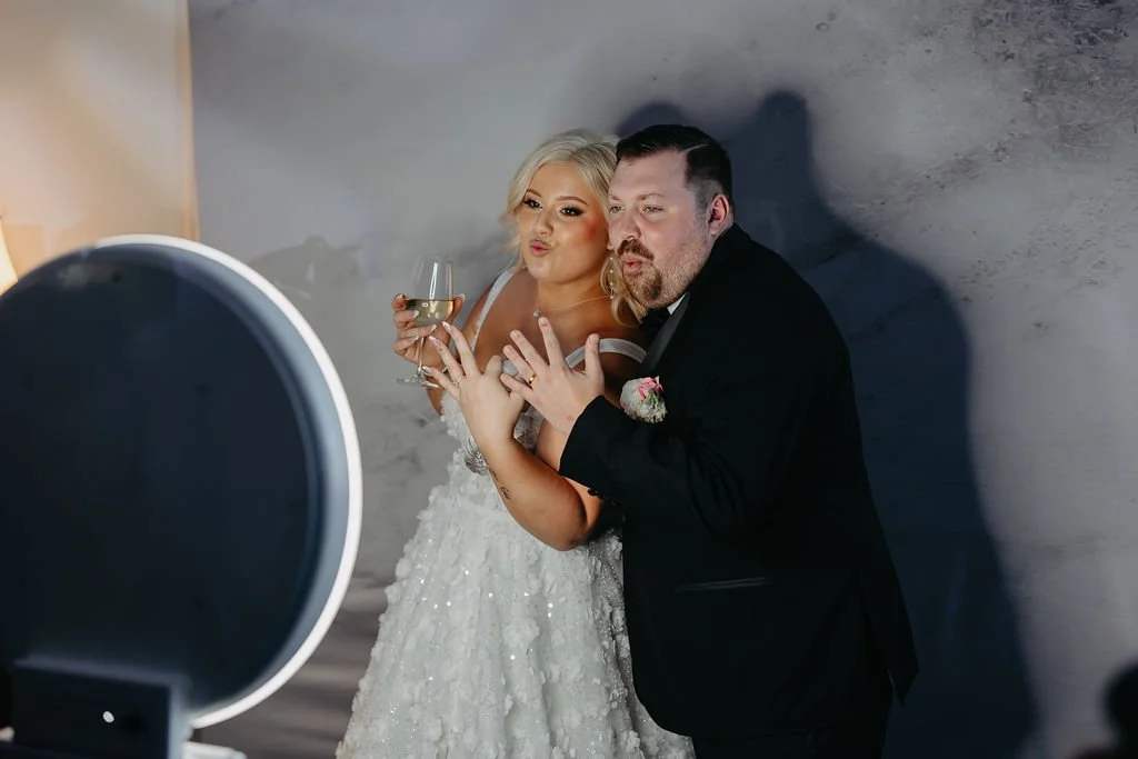 A bride and groom taking selfies together while holding a glass of wine, celebrating at their wedding.