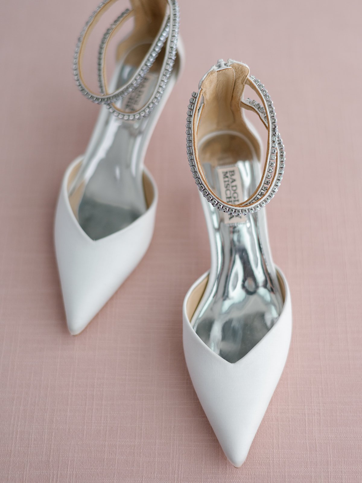 A pair of white high-heeled shoes with pointed toes and ankle straps adorned with rhinestones, placed on a pink surface.