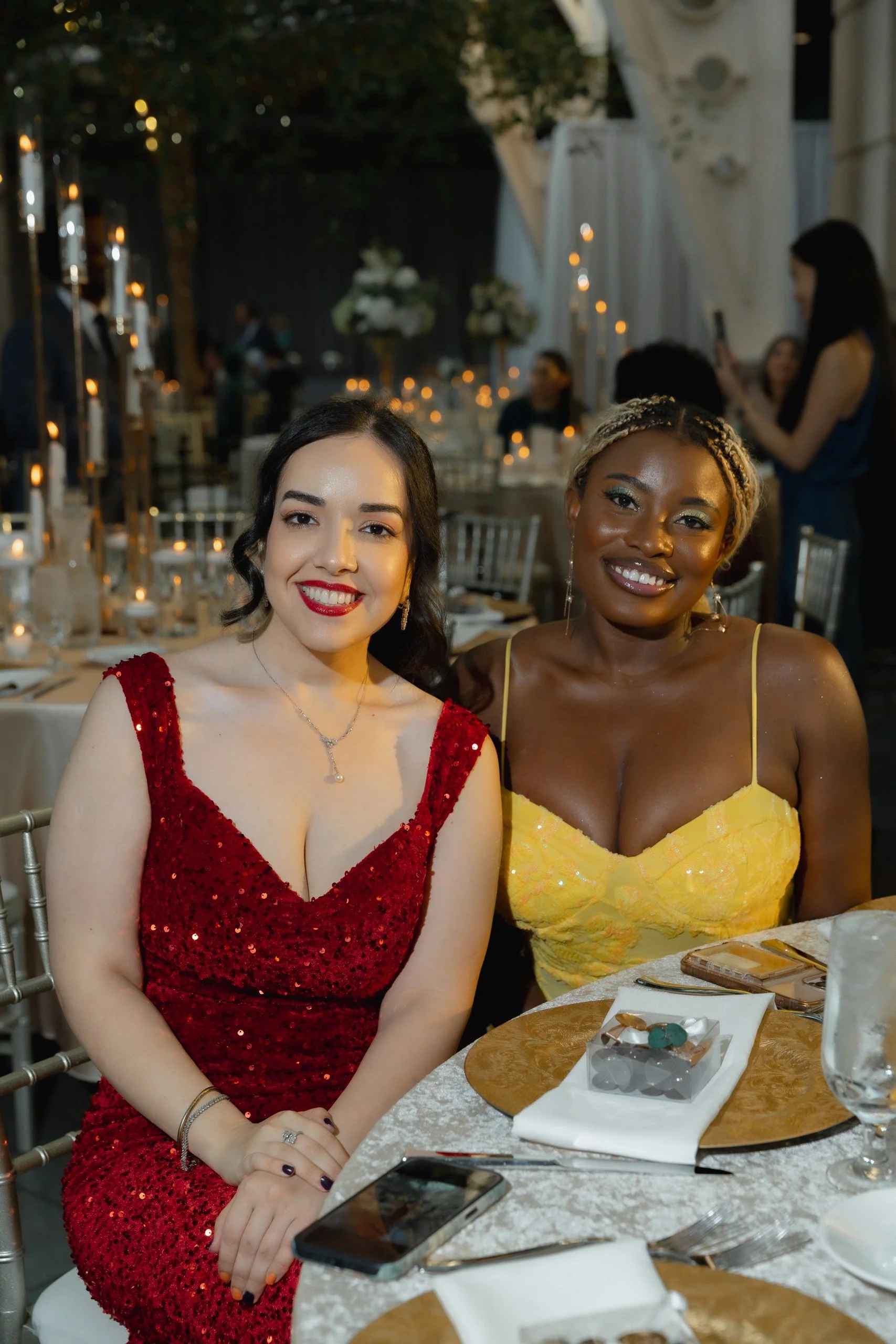 Two women sitting at a dinner table during an evening event, smiling at the camera. One woman is wearing a red sequined dress, and the other is wearing a yellow dress with spaghetti straps. The table has a white tablecloth, plates, silverware, a glas