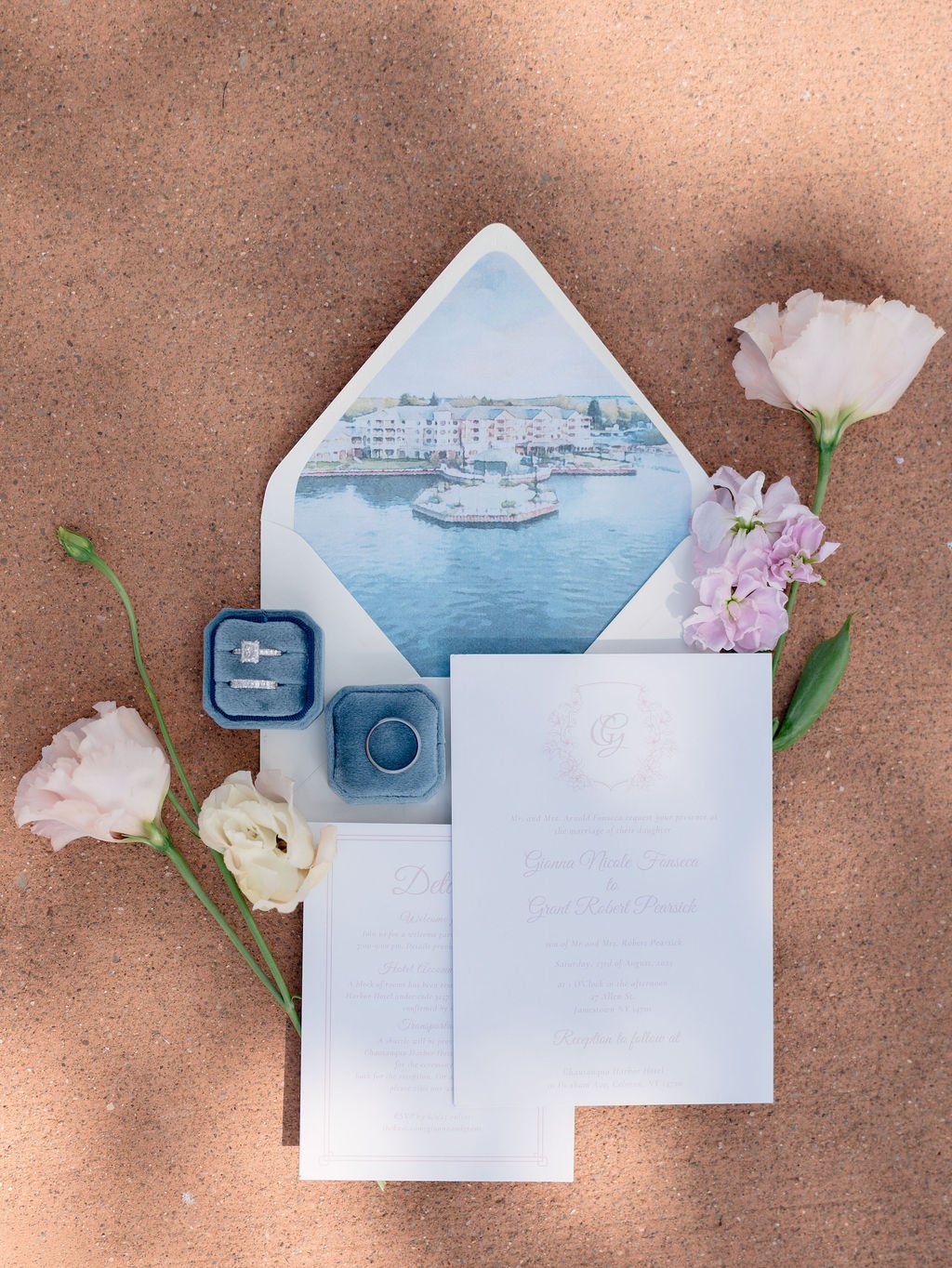 Wedding invitation with floral decorations, engagement ring, wedding bands, and a waterfront scene in the background.