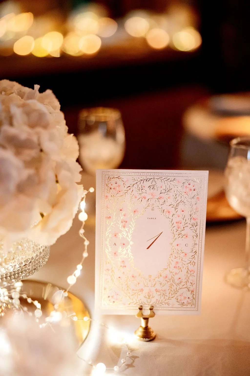 Elegant wedding reception table with floral centerpiece, table number card, and fairy lights in warm lighting.