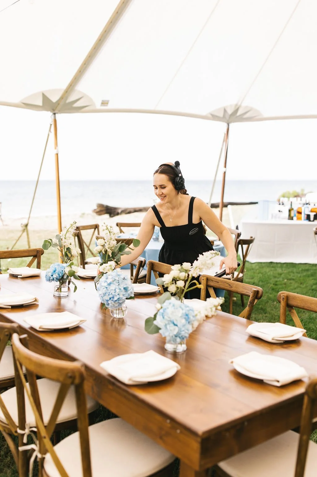A woman wearing a black dress and headphones setting up a long wooden table with floral centerpieces at an outdoor event near the beach, under a large white canopy.