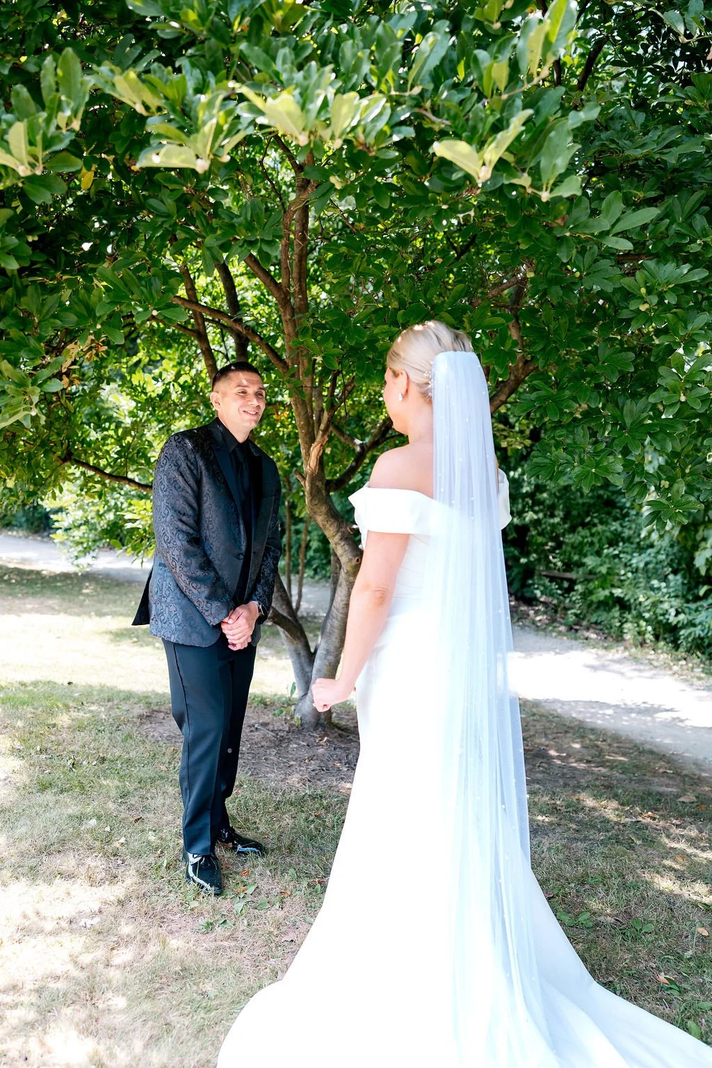 A bride and groom standing outdoors under a lush green tree, facing each other during their wedding ceremony.