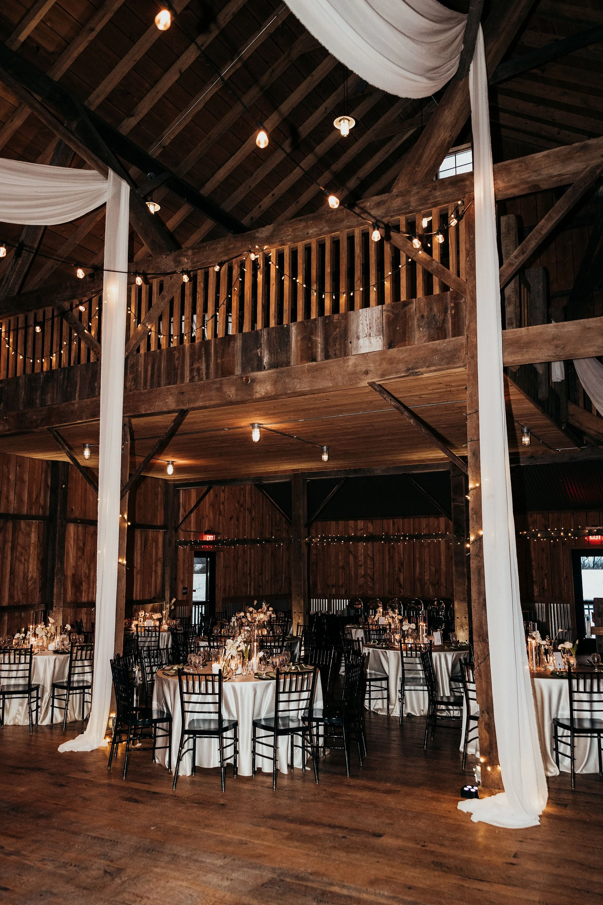 Inside a rustic barn decorated for a wedding reception with string lights, round tables covered with white tablecloths, and black chairs. White drapes hanging from the upper level, warm lighting, wooden beams, and flooring.