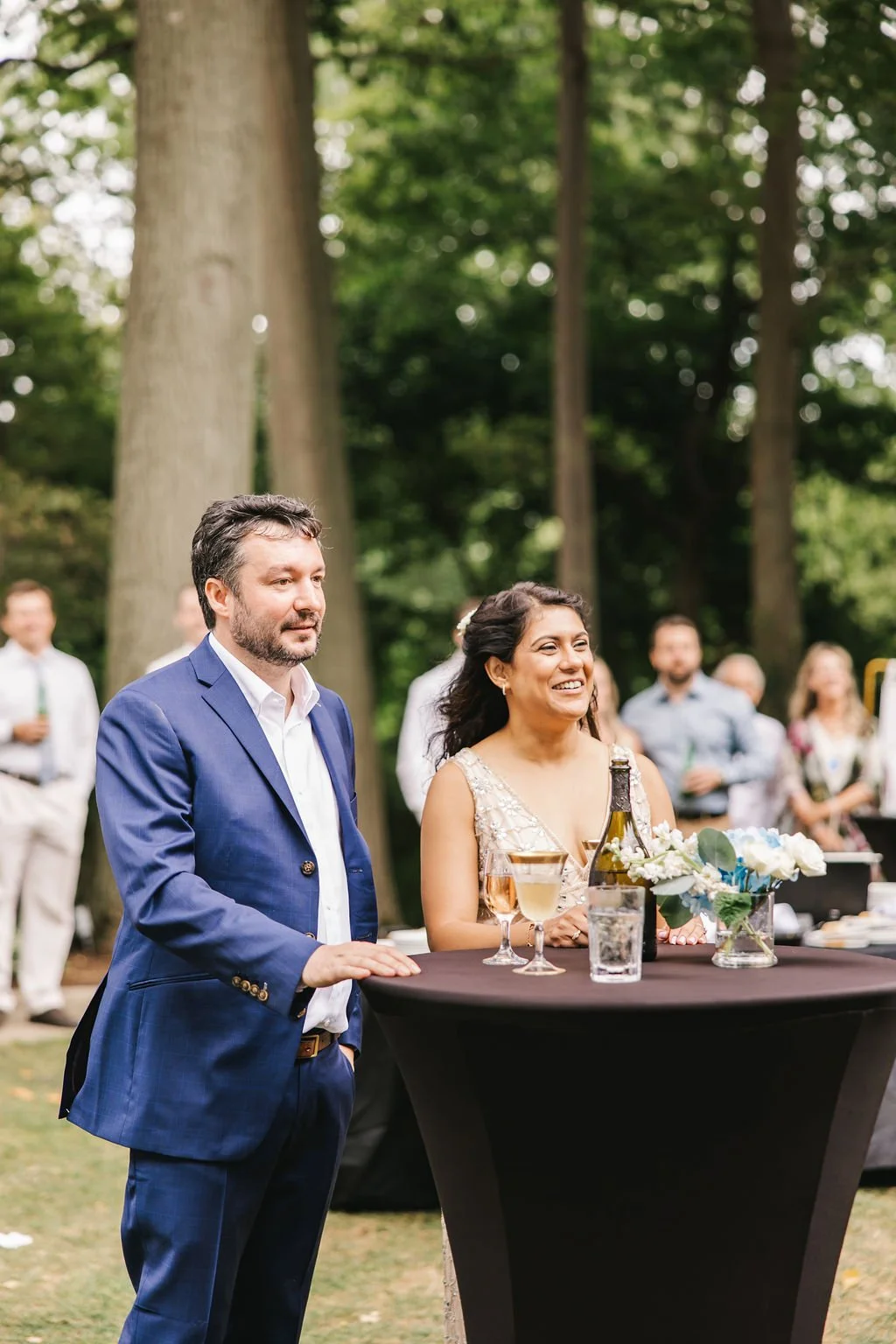 A man in a blue suit and a woman in a beige dress standing at a black cocktail table outdoors during a celebration, surrounded by trees and other people in the background, with drinks and floral decor on the table.
