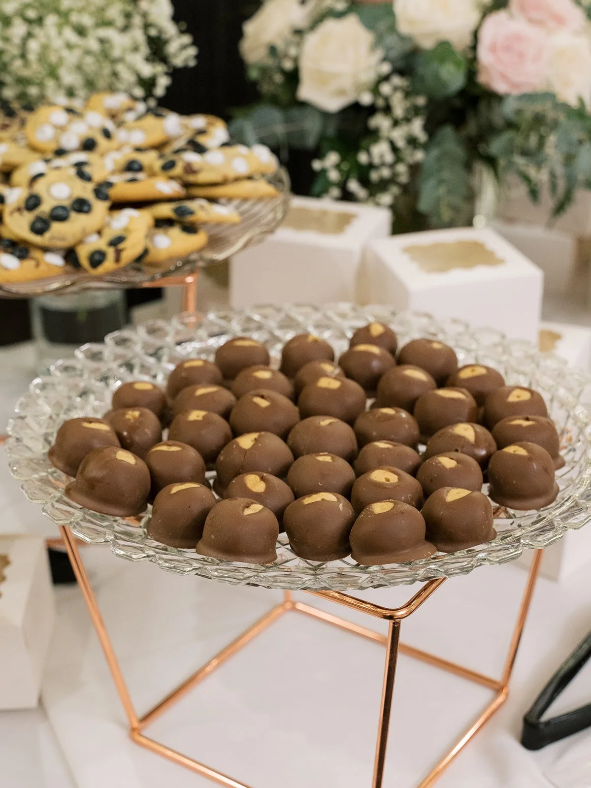 Chocolate-covered candies with white filling on a glass tray, with cookies and flowers in the background.