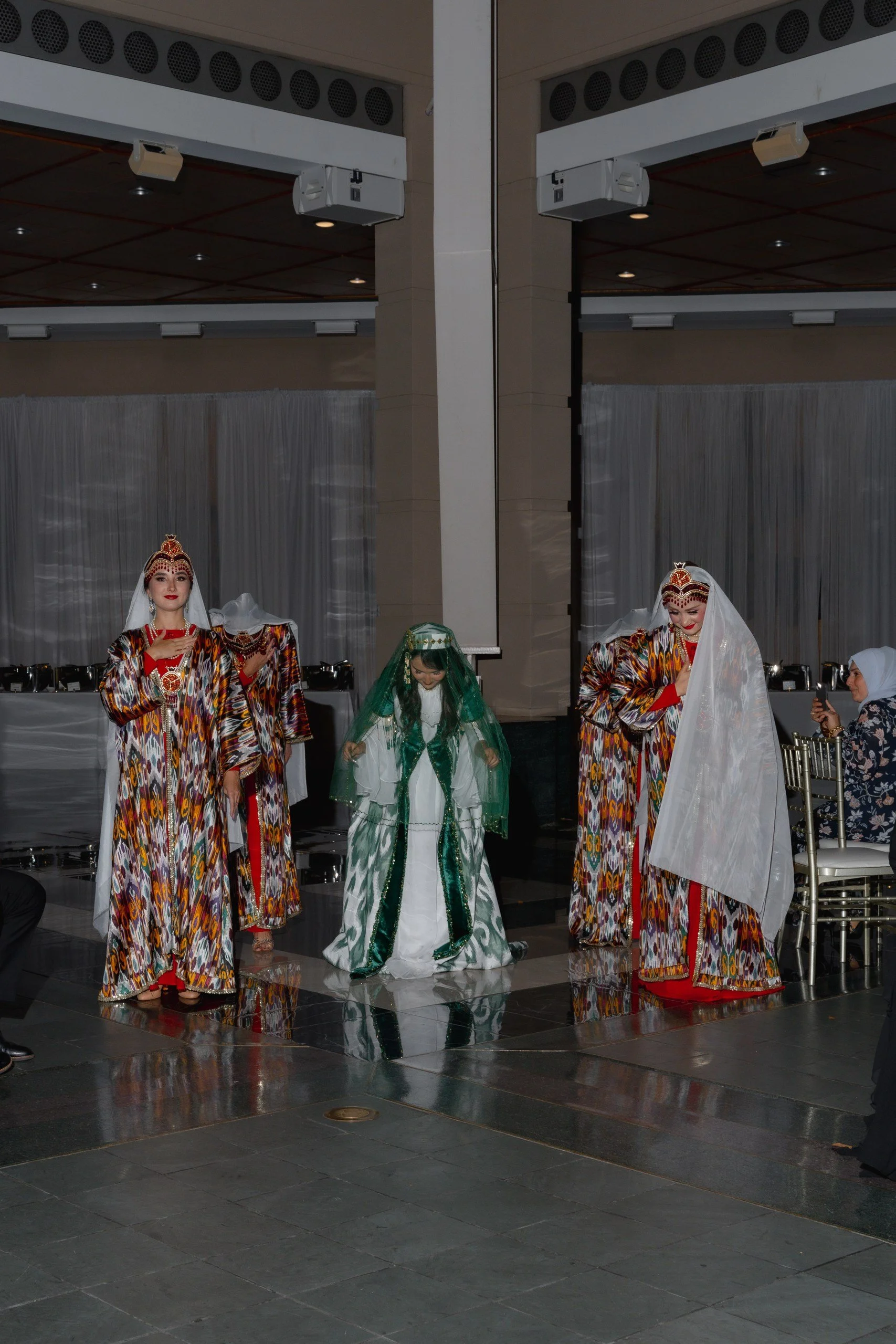 Two women dressed in traditional colorful dresses are participating in a cultural dance or celebration indoors. They are wearing elaborate headpieces and jewelry, with one holding a white veil. There are tables with covered dishes and a woman in mode