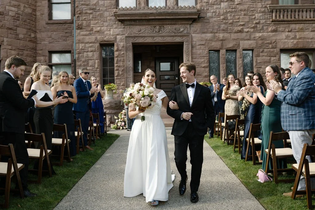 Bride and groom walking down the aisle at a wedding ceremony, surrounded by guests clapping and smiling outside a stone building.