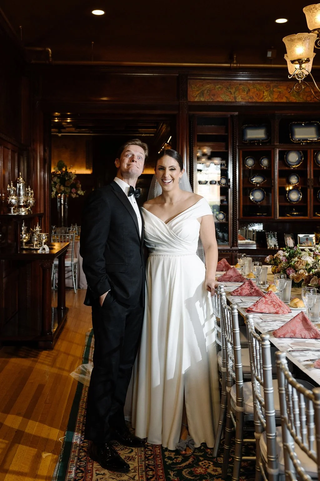 A bride and groom in wedding attire standing inside a decorated banquet room, smiling at the camera.