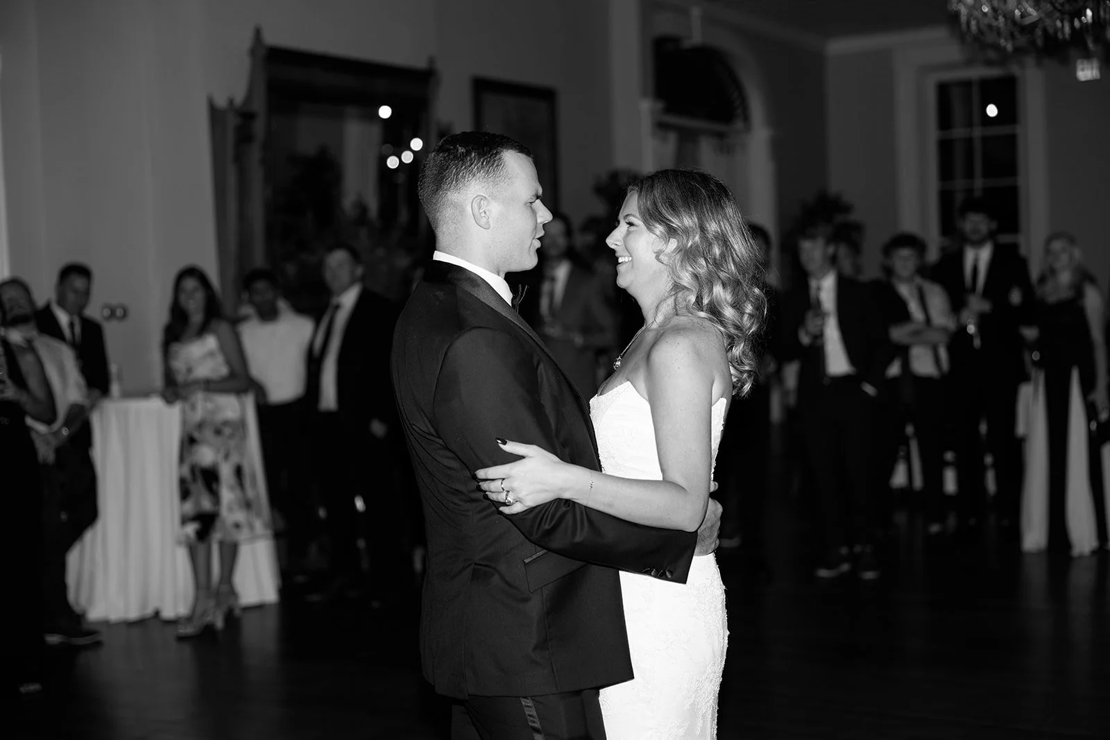 A bride and groom dancing at their wedding reception, smiling at each other, while guests watch in the background.