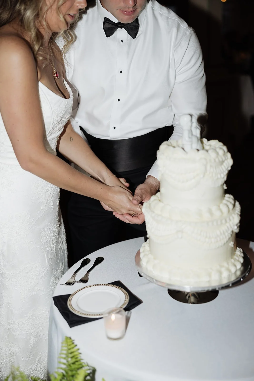 A bride and groom, dressed in wedding attire, holding hands over a wedding cake, preparing to cut it at their wedding celebration.
