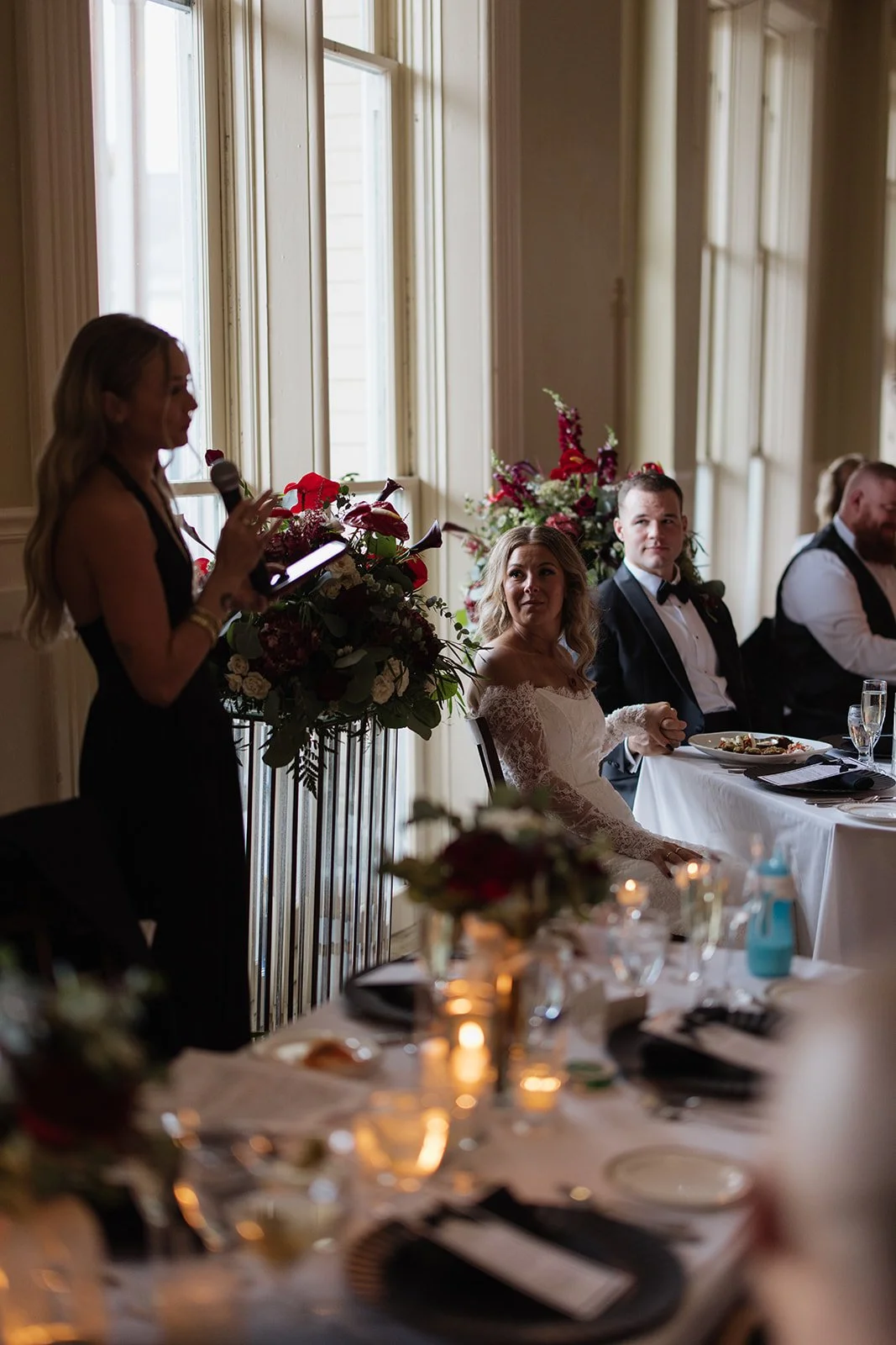 A woman giving a speech at a wedding reception, with guests seated at tables decorated with flowers and candles, in a room with large windows and natural light.