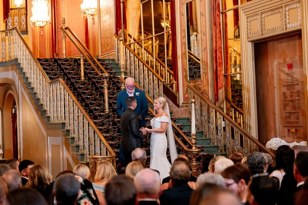 A wedding ceremony taking place on a staircase inside a grand, ornate building with gold accents and red-orange wallpaper. The bride and groom are exchanging vows with an officiant standing behind them. Guests are seated at the bottom of the staircas