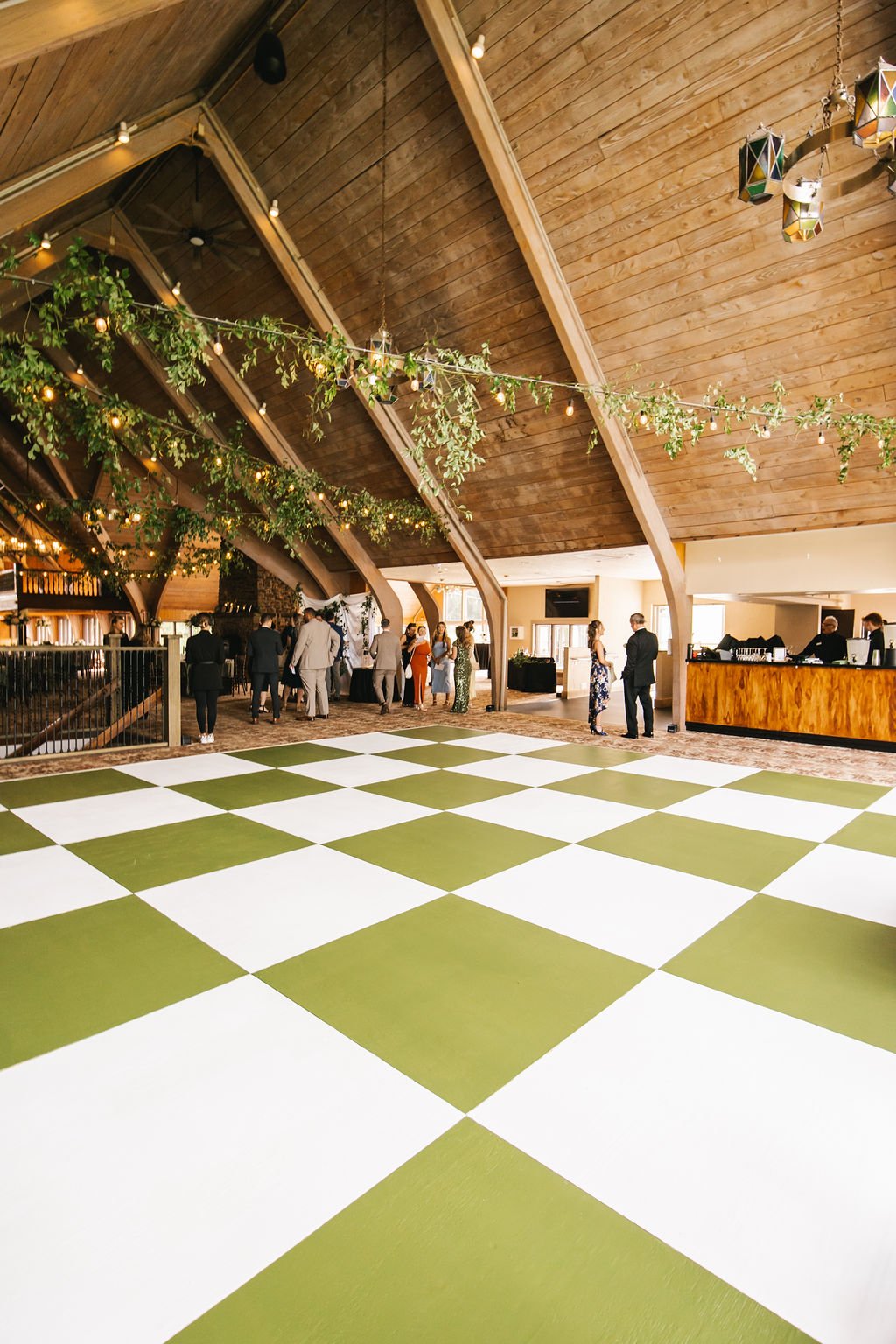 Indoor wedding venue with wooden ceiling, hanging string lights, green foliage, and a checkered green and white dance floor, with guests mingling near the bar.