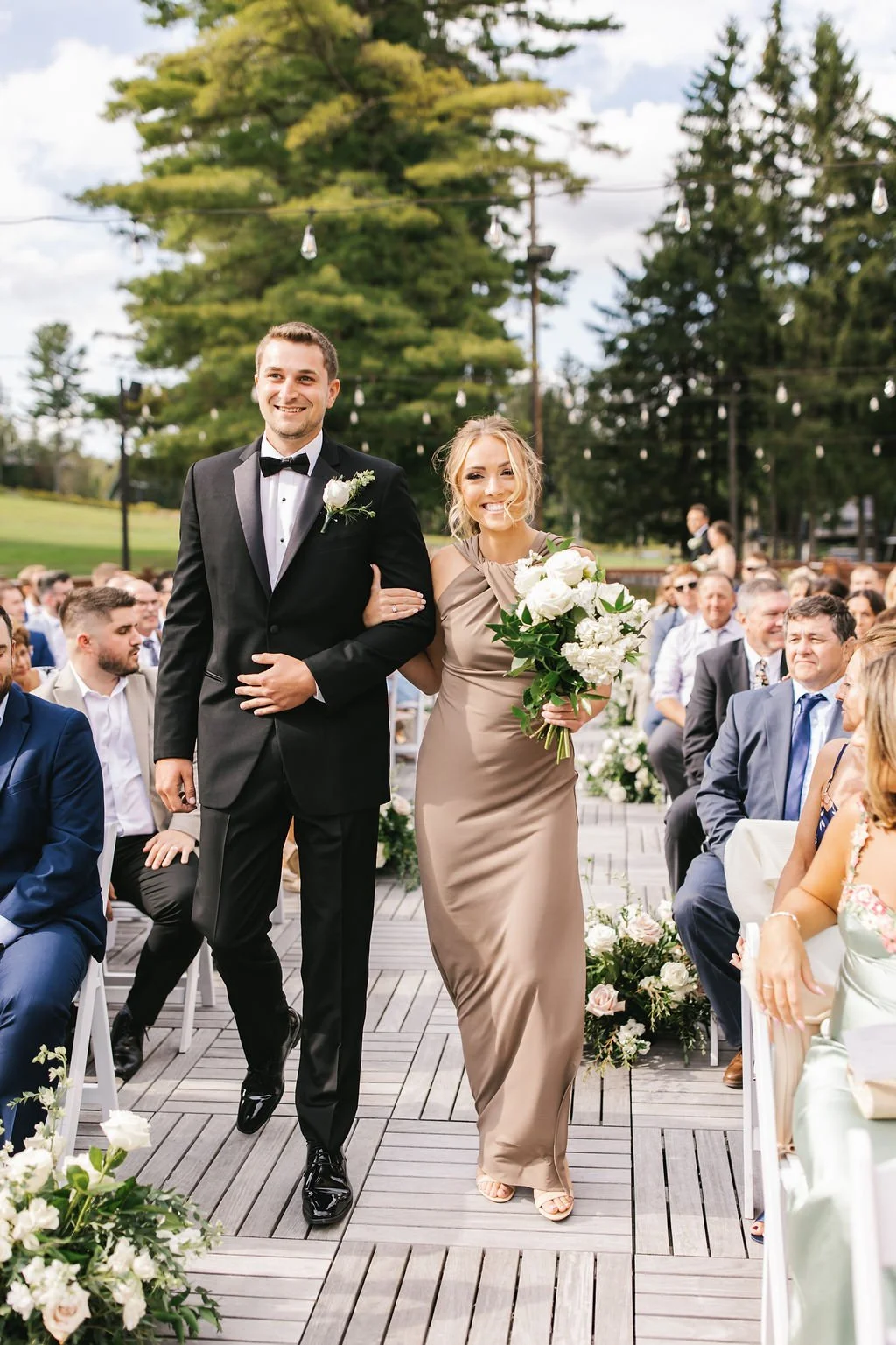 A bride and groom walking down the aisle at an outdoor wedding ceremony, surrounded by seated guests and decorated with flowers and greenery.