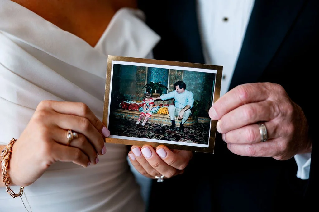 Two people holding a framed photo of a man and a young girl sitting on a decorative floor with flowers around them.