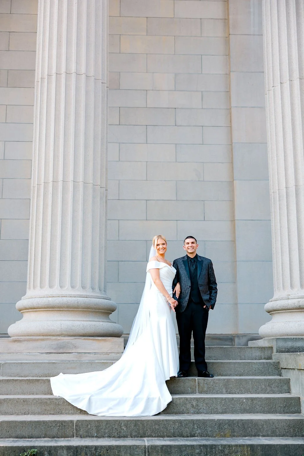 A bride and groom stand on steps in front of large stone columns and a stone wall, smiling and holding hands.