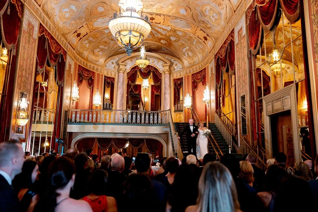 A bride and groom walking down the stairs at a wedding reception inside an opulent, gold-accented hall with chandeliers and mirrors, surrounded by seated guests.