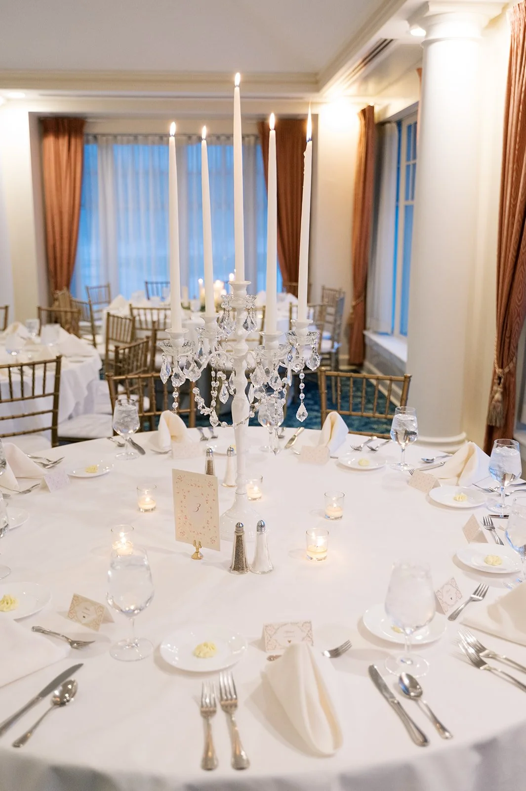 Elegant banquet table setup with white tablecloth, silverware, glassware, candles, and a crystal chandelier centerpiece in a decorated room with large windows and draped curtains.