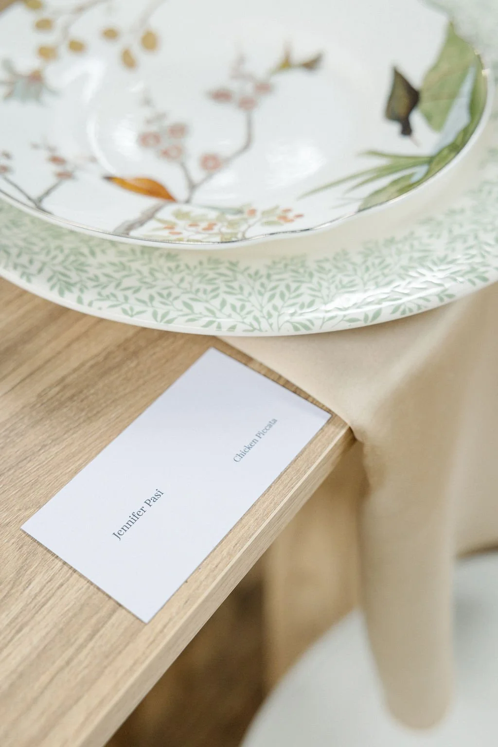 A decorative plate with floral and leaf designs placed on a wooden table, with a white card labeled 'Jennifer Pasi' and 'Chicken Pasta' next to it.