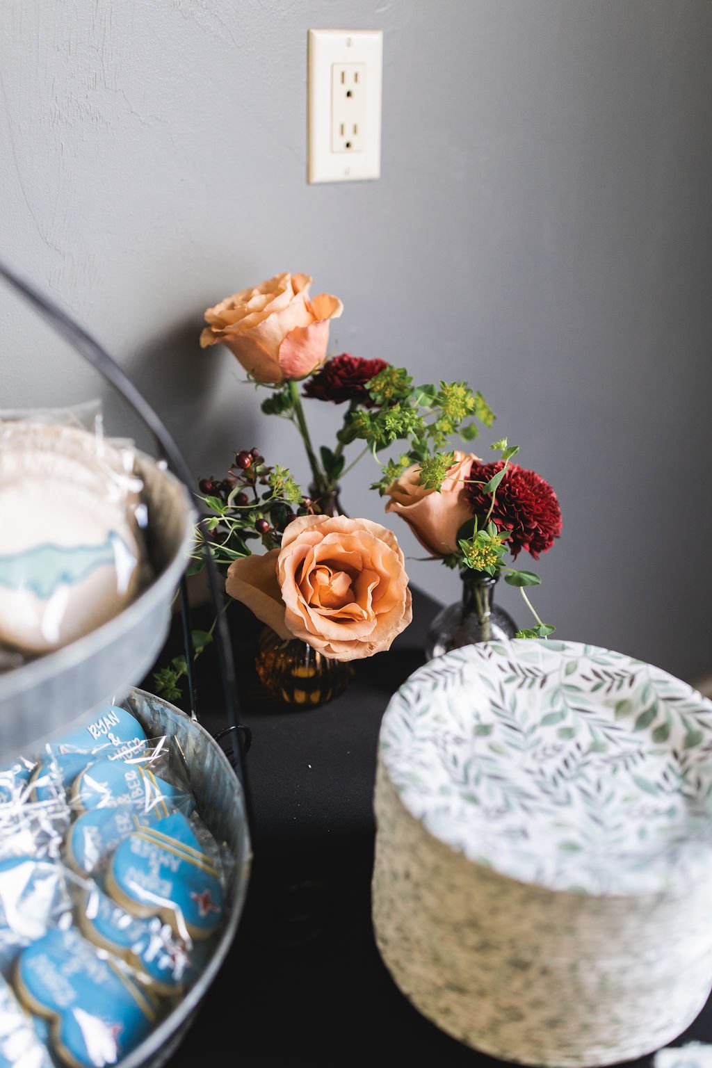 A table with three small vases of peach and red flowers, a decorative paper-tuff rice bowl, and a container of wrapped candies. There is a gray wall with an electrical outlet in the background.