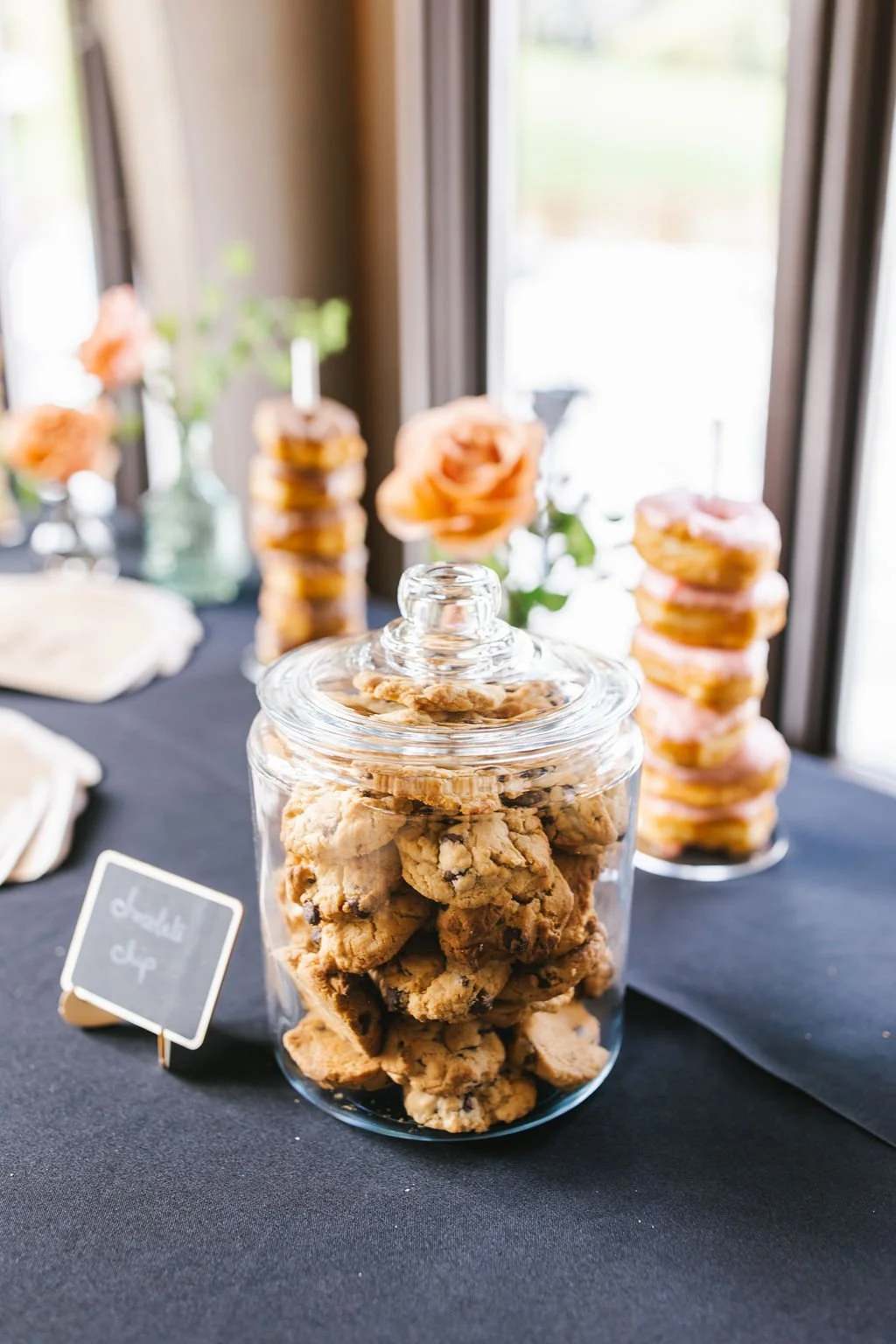 Glass jar filled with chocolate chip cookies on black table at a dessert table, with donuts on a stand and flowers in the background.