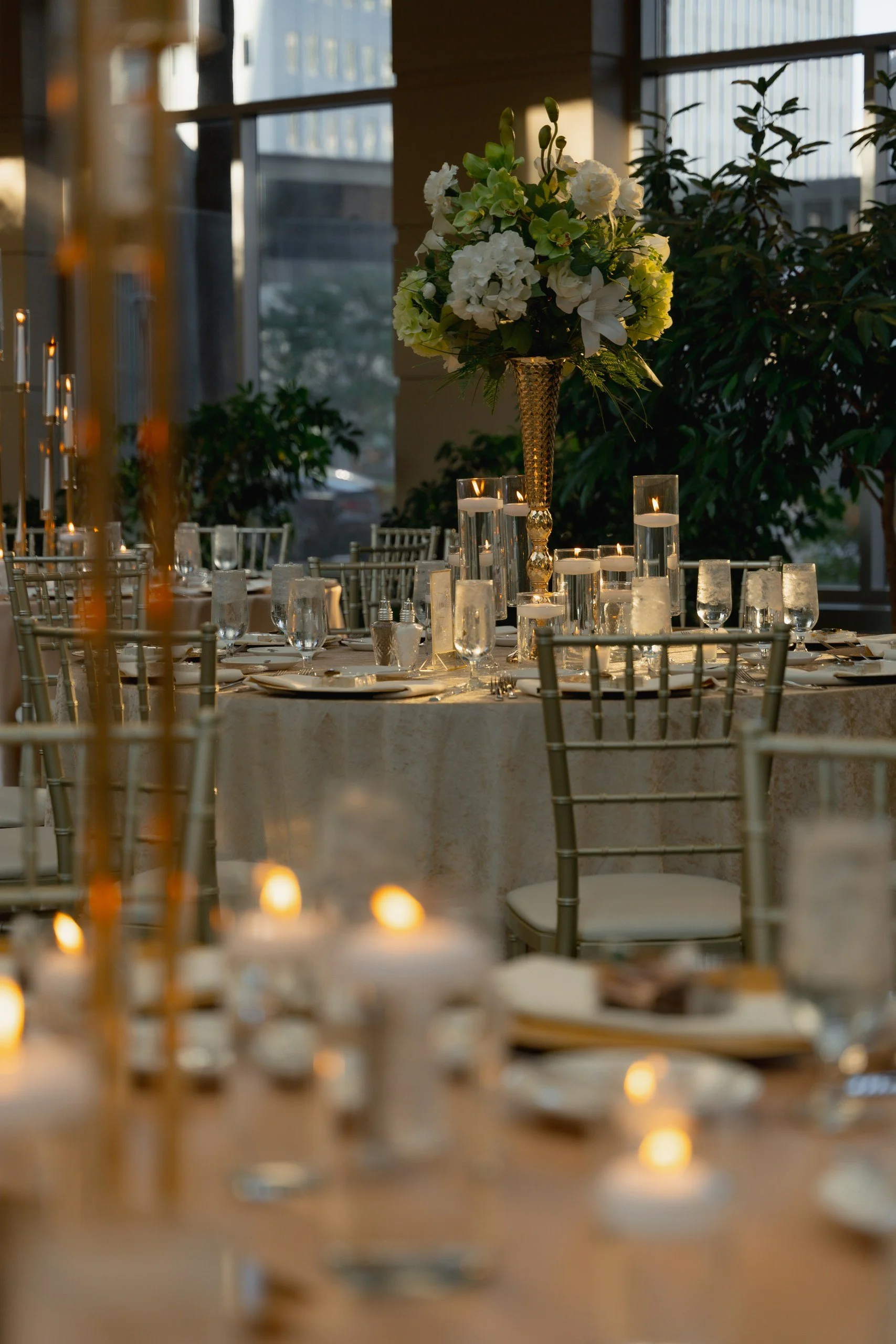 Elegant event table with floral centerpiece in a tall gold vase, surrounded by lit candles and glassware, set in a well-lit room with large windows and greenery in the background.