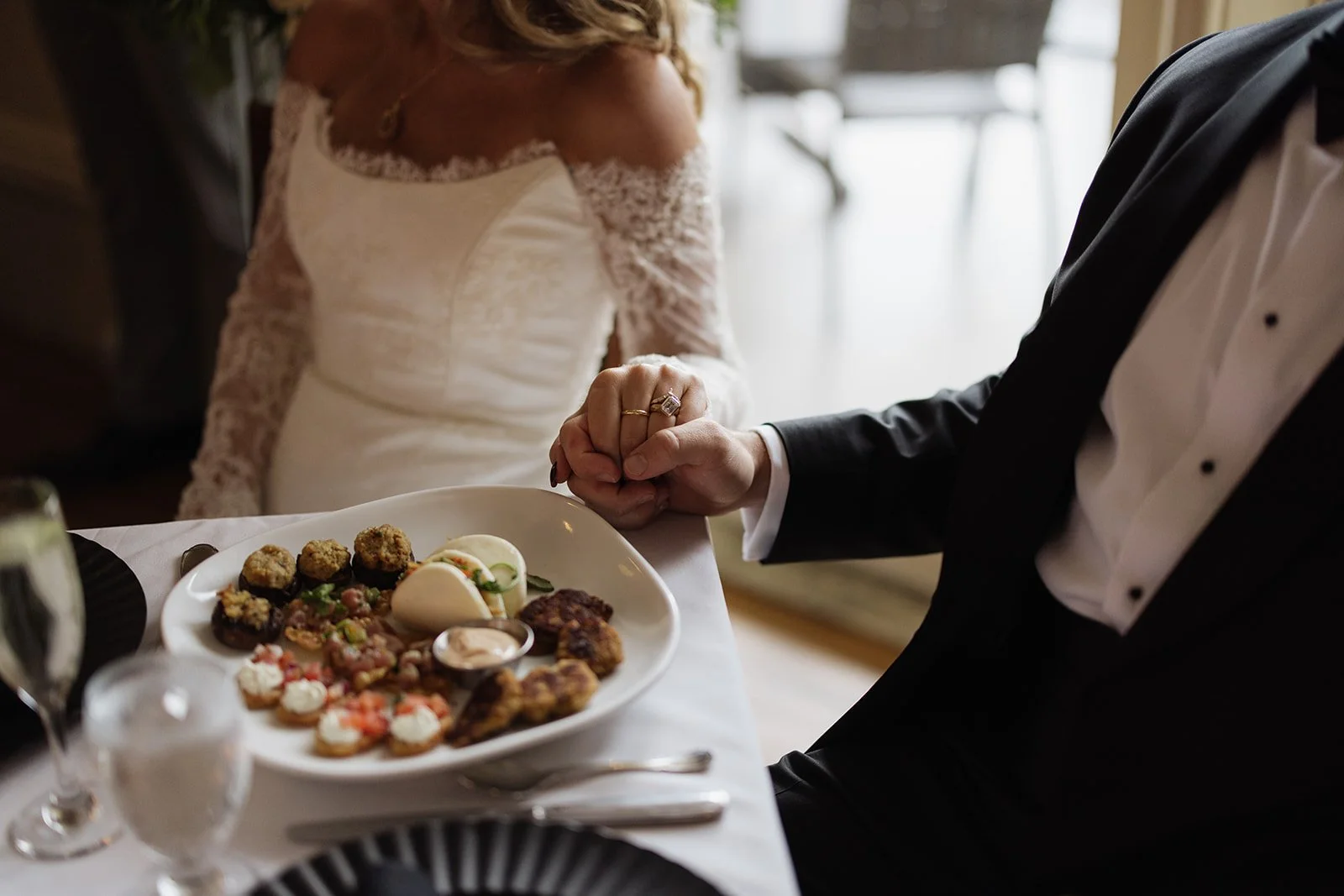 A bride and groom holding hands at a wedding reception table, with the bride wearing a lace wedding dress and the groom in a black tuxedo. The table has a platter of various appetizers and finger foods.