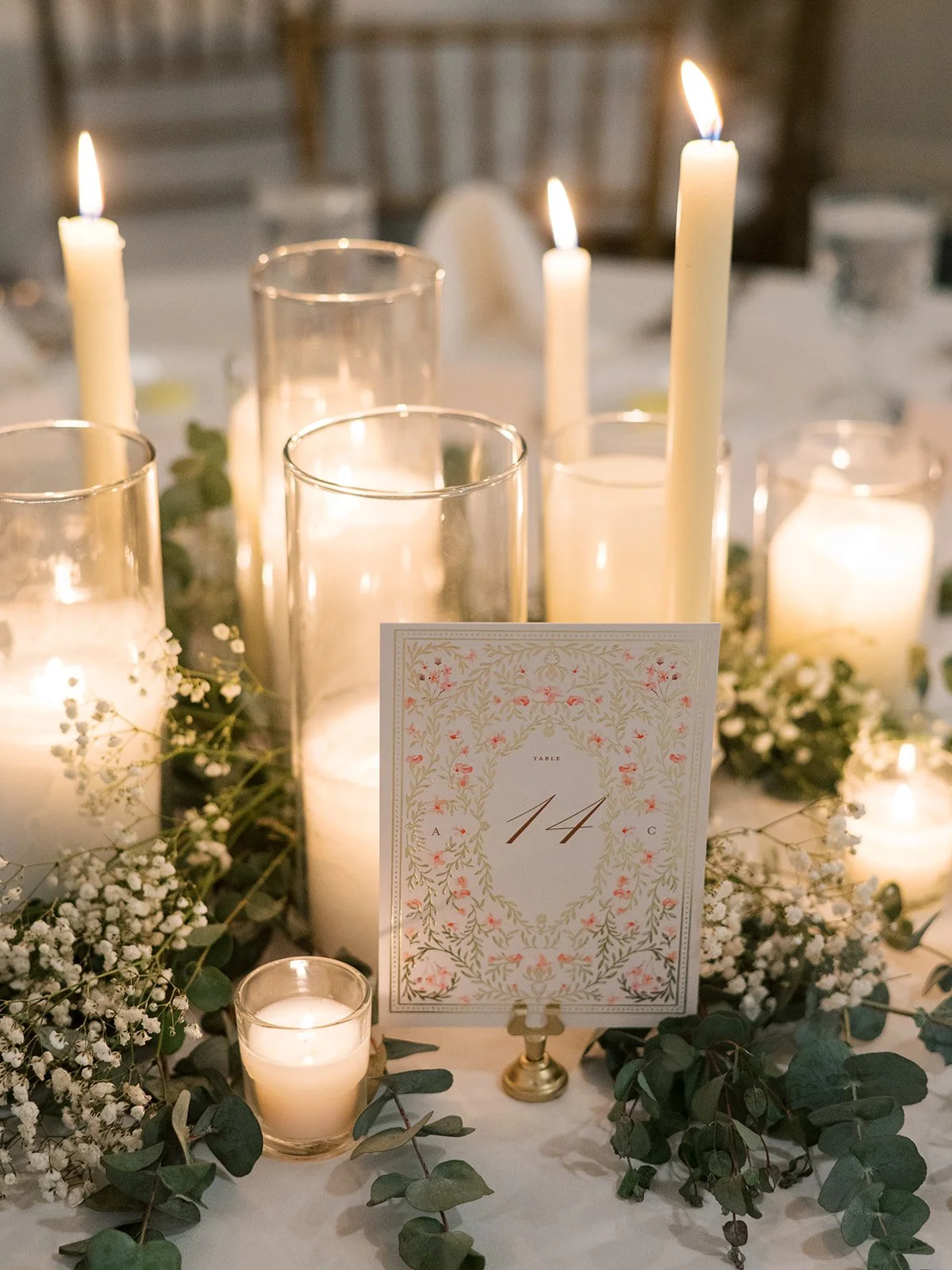 Elegant table centerpiece with tall white candles in glass holders, surrounded by greenery and white flowers, with a table number card displaying '14' in a decorative frame.