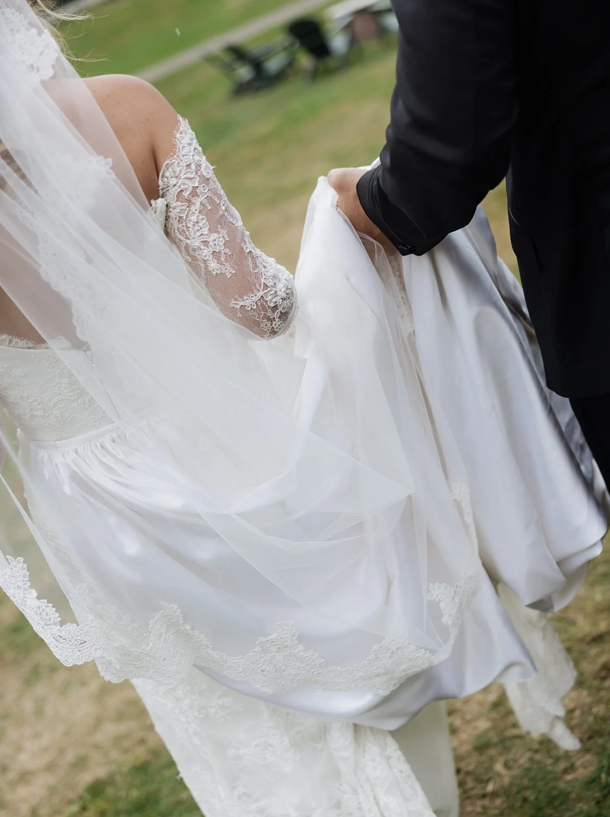 A bride wearing a lace wedding dress lifting her wedding gown with the help of the groom dressed in a black suit, outdoors on grass