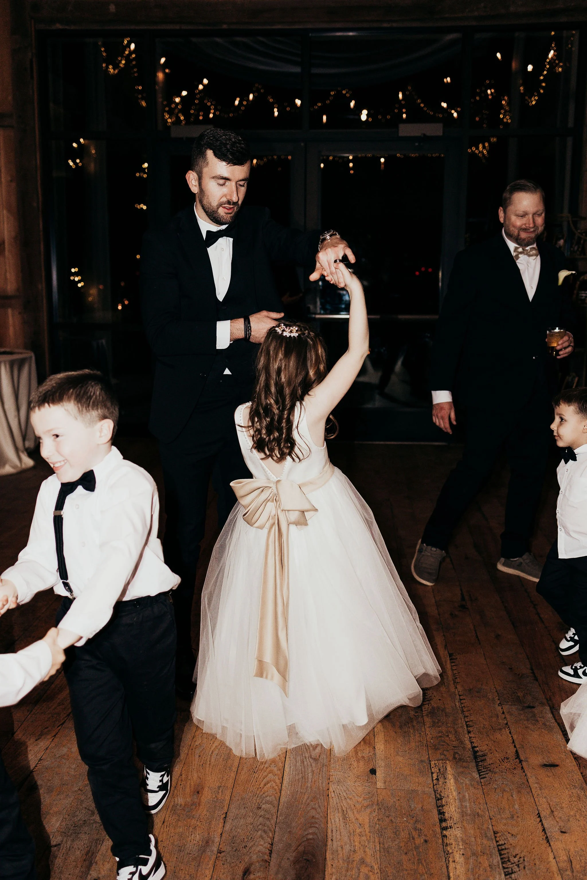 Children and adults dancing at a wedding reception with wooden floors, string lights, and large windows in the background.