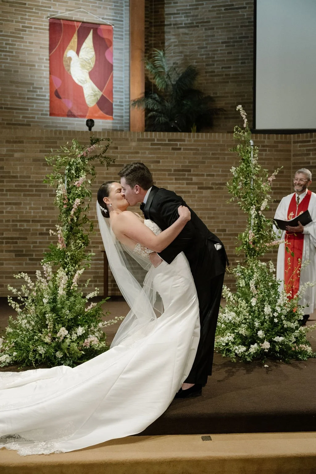 A wedding couple sharing a kiss during their ceremony, with the bride in a white gown and veil and the groom in a black tuxedo, in front of floral arrangements. An officiant stands in the background smiling.
