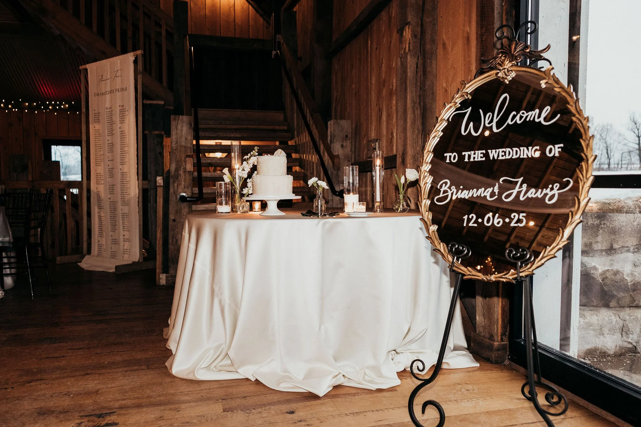 Decorative wedding sign with gold frame reading 'Welcome to the wedding of Brianna & Travis 12-06-25', placed near a table with a white wedding cake decorated with white flowers and candles, inside a rustic wooden venue.
